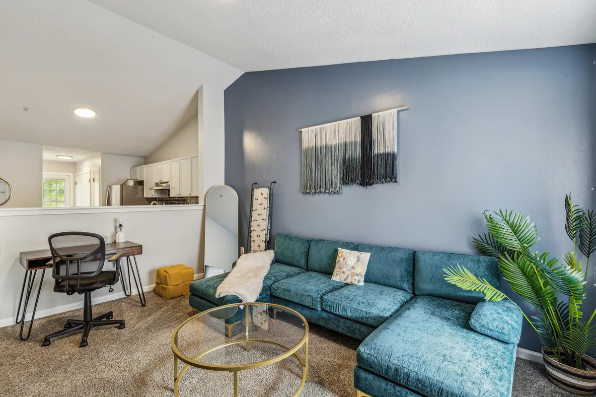 A modern living area with a teal sectional sofa, glass coffee table, decorative wall hanging, and lush potted plant. The room also features a workspace with a chair, offering flexibility for leisure or remote work. The stylish, open design connects seamlessly with other parts of the home, emphasizing communal living.
