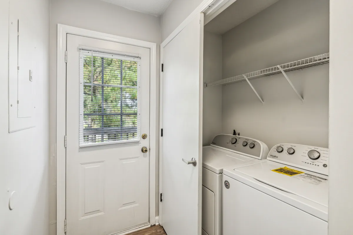 This image presents a bright and functional laundry area, featuring a modern washing machine and dryer set next to an entry door and a window that lets in natural sunlight. The practical layout shows the space's utility while the sunlight adds a touch of warmth to the room.