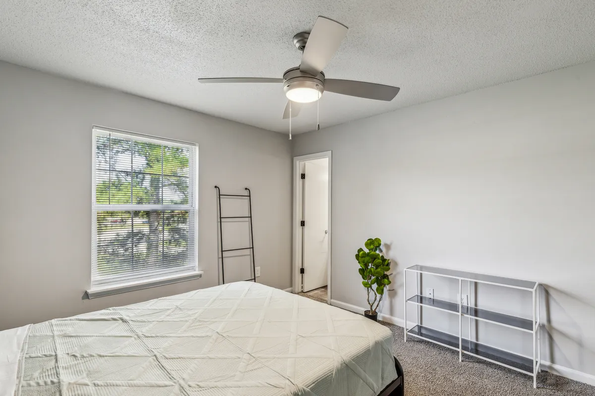 A tranquil bedroom scene featuring a neatly made bed beside a sunlit window, a small plant adding a touch of greenery, and a tidy shelf. The space feels calm and welcoming, ideal for relaxing, reading, or catching up on work in peace.