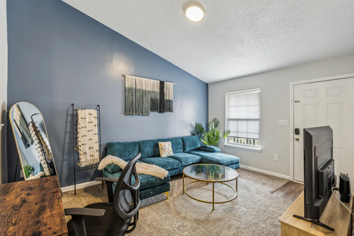 A spacious living room featuring a striking teal sectional sofa set around a round glass coffee table. Decorative wall art and a large mirror add contemporary flair, while a potted plant introduces a touch of nature. The room exudes a cozy, modern vibe and is flooded with natural light through a window, inviting moments of relaxation or socializing.
