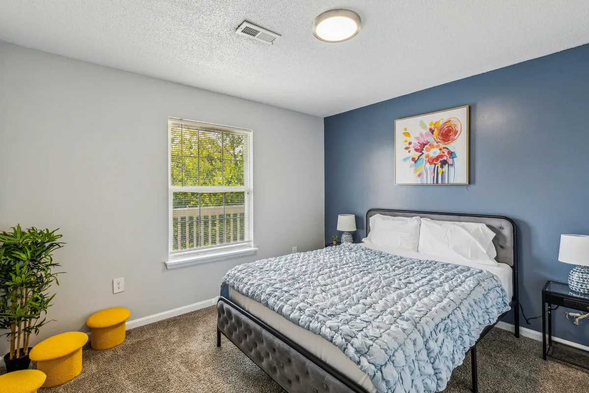 A bright, serene bedroom featuring a queen bed with a blue quilt, floral artwork above the bed, yellow stools at the foot, a large window letting in natural light, and a potted plant adding a fresh touch.