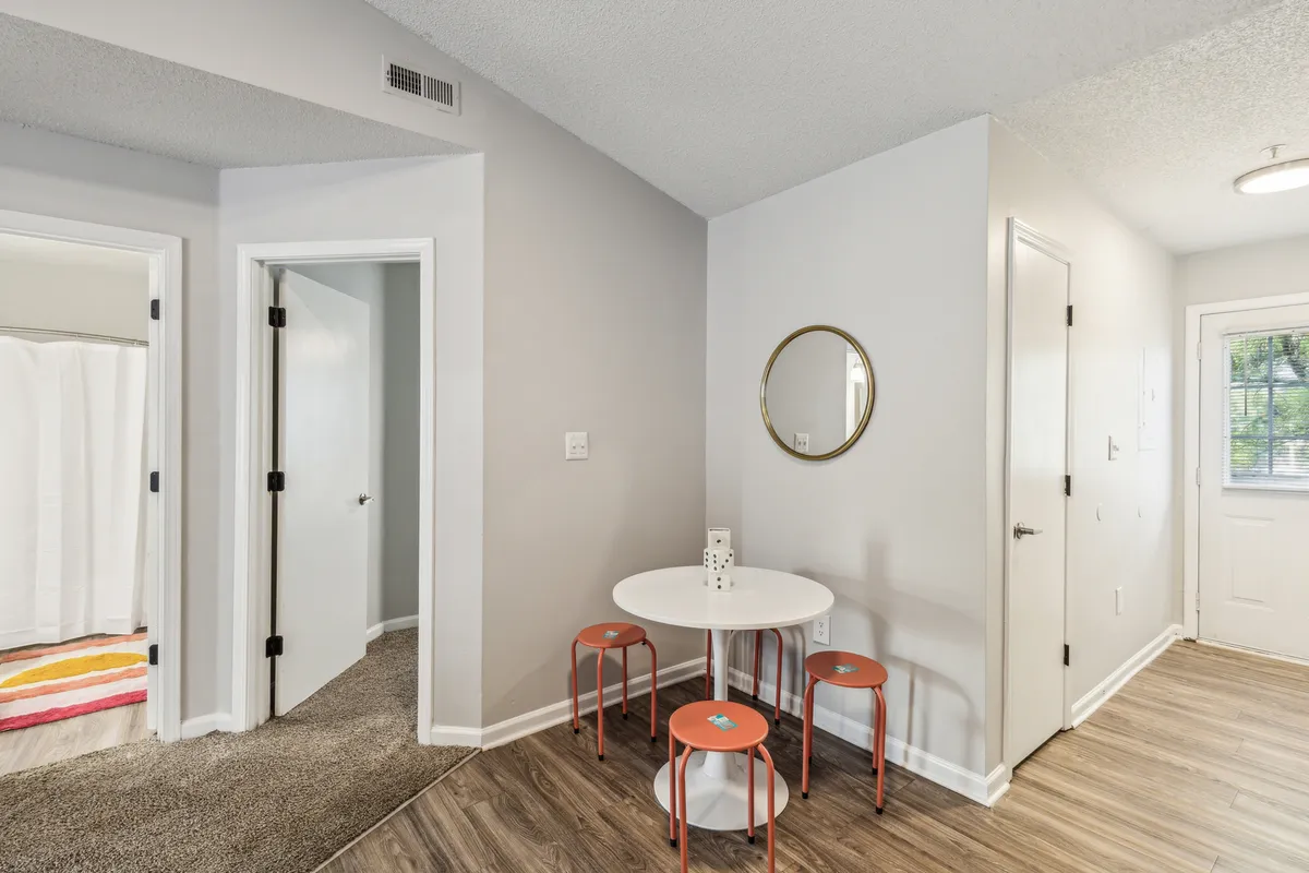 A cozy dining nook featuring a small round table with orange chairs and a mirror, set in a welcoming, softly lit area. The setup is perfect for casual dining, intimate conversations, or a friendly game night, complementing the kitchen area.