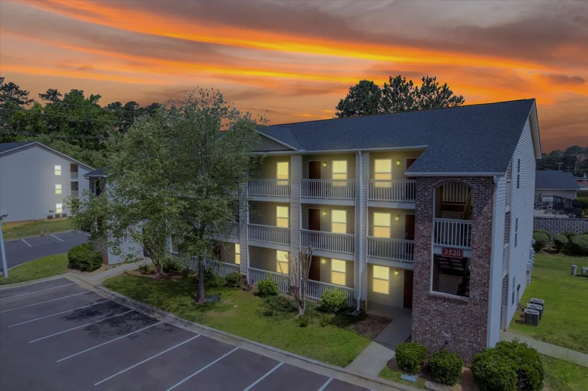 A three-story apartment building is shown with balconies overlooking green spaces, set against a serene sunset sky. Residents can be imagined relaxing on balconies or engaging in social interactions in the courtyard area below.