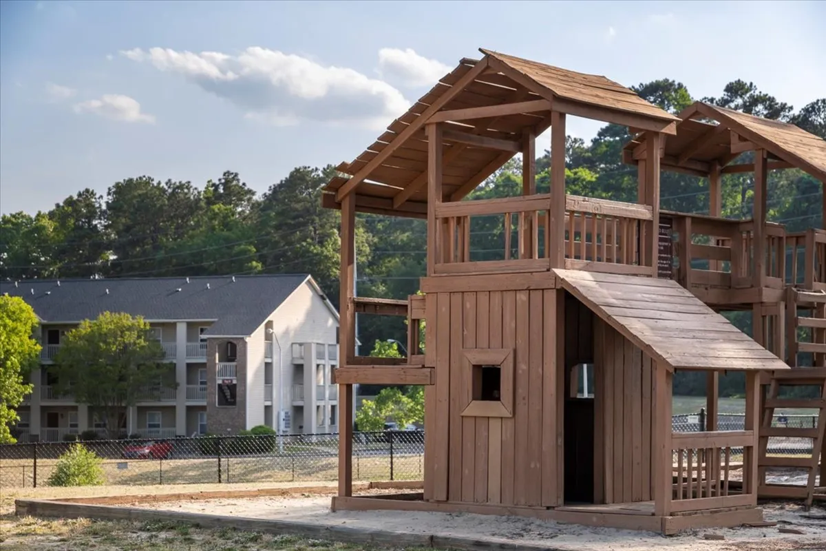 The image captures a vibrant playground structure surrounded by lush greenery with an apartment building visible in the background. Children are seen playing, and there's a sense of community and family-friendly atmosphere outdoors.