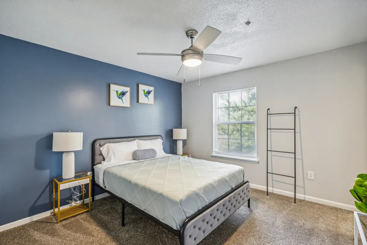 A calm bedroom highlighted by a blue accent wall, a queen bed with soft linens, bedside lamp, window bringing in natural light, and tasteful wall art, creating a soothing, restful space.